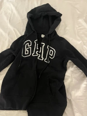 Svart GAP hoodie med dragkedja - Står s men den är mer xxs för den sitter tight,Svart hoodie från GAP med vit broderad logga på bröstet. Tröjan har dragkedja framtill, två fickor och justerbar huva med snören. Perfekt för en avslappnad och cool stil.