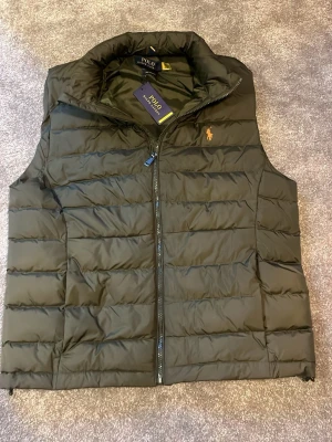 Olivgrön dunväst från Polo Ralph Lauren - Snygg olivgrön dunväst från Polo Ralph Lauren med orange broderad logga på bröstet. Västen har hög krage, dragkedja framtill och två sidofickor. Tillverkad i ett glansigt material som ger en modern look. Perfekt för lager-på-lager-stil. (Rep)
