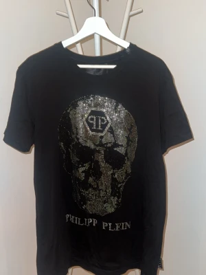 Svart Philipp Plein t-shirt med paljetter - Snygg svart t-shirt från Philipp Plein med korta ärmar och rund hals. Framsidan har ett stort motiv i glittriga paljetter och logga, medan baksidan har en liten metallpatch vid nacken. Perfekt för dig som gillar statement-plagg.