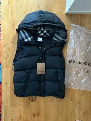 Burberry jacka - Snygg svart dunväst från Burberry med avtagbar huva och klassiskt rutigt foder. Västen har dragkedja och knappar framtill, två fickor med dragkedja och logga på bröstet. Perfekt för lager på lager och streetstyle.