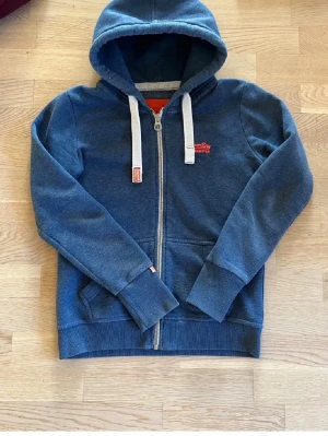 Blå zip hoodie från Superdry - Mörkblå hoodie från Superdry med dragkedja framtill, vita snören i huvan och fickor på sidorna. Liten Superdry-logga i rött på bröstet och en etikett på ärmen. Mjuk bomullsmix och ribbade muddar vid ärmslut och nederkant.