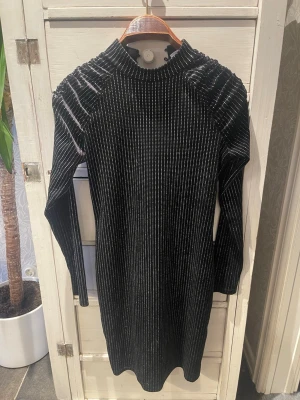 Svart glittrig kortklänning - Snygg svart kortklänning med glittriga ränder och hög krage från Puls Kolm. Klänningen har långa ärmar, markerade axlar och dragkedja i ryggen. Perfekt för dig som vill sticka ut med en trendig och elegant look.