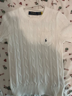 Vit kabelstickad tröja Polo Ralph Lauren - Kabelstickad vit tröja från Polo Ralph Lauren med korta ärmar och rund hals. Tröjan har en liten bröstficka med den klassiska broderade loggan i marinblått. Ribbstickade muddar vid ärmslut och nederkant. Perfekt för en clean och stilren look.