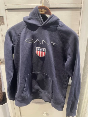 Mörkblå hoodie från GANT - Mörkblå hoodie från GANT med broderad logga och amerikanskinspirerat sköldmärke på bröstet. Klassisk känguruficka framtill och stor huva. Tillverkad i mjukt bomullsmaterial, perfekt för en avslappnad och stilren look.