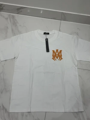 Vit Amiri t-shirt med logga - Snygg vit t-shirt från Amiri med korta ärmar och gulbrun logga på bröstet samt stort AMIRI-tryck på ryggen. Tillverkad i mjuk bomull och har en klassisk passform. Perfekt för dig som gillar stilrena plagg med exklusiv känsla.