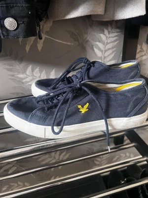 Marinblå sneakers från Lyle & Scott - Snygga marinblå sneakers från Lyle & Scott med klassisk låg profil och vit sula. Skorna har gul broderad logga på sidan och är gjorda i canvasmaterial. Perfekta för en avslappnad och stilren look.