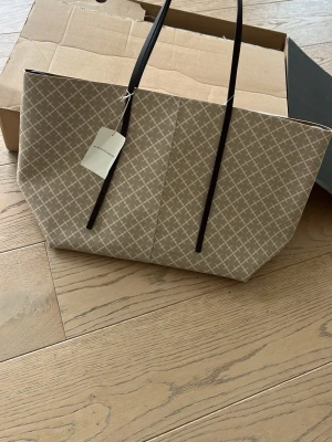ABI  TOTE Beige shoppingväska från By Malene Birger - Stilren shoppingväska i beige med vitt grafiskt mönster från By Malene Birger. Väskan har svarta långa axelband och rymligt fack med matchande innerficka. Tillverkad i tåligt konstläder som ger en lyxig känsla. Perfekt för dig som gillar trendiga accessoarer.