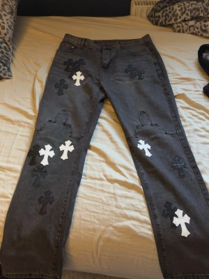 Chrome Hearts Jeans - Unika grå jeans med broderade svarta och vita korsapplikationer på både fram- och baksida. Rak passform och klassisk femficksdesign. Jeansen har en edgy streetwear-vibe och är perfekta för dig som vill sticka ut. Materialet är denim i bomull.