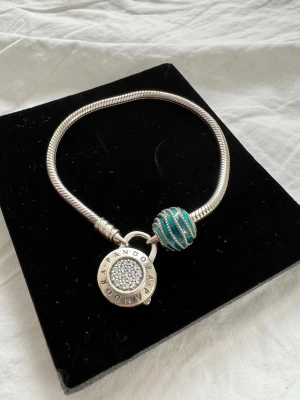Pandora silverarmband med pärldetalj - Snyggt silverfärgat armband från Pandora med en rund berlock och en blågrön pärla med glittrande detaljer. Armbandet har en klassisk orm-länk och berlocken är dekorerad med små stenar och Pandora-logga. Box finns inte kvar! 