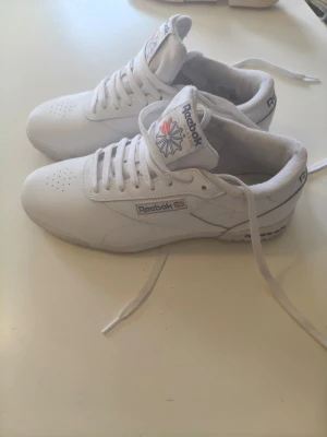 Vita Reebok Classic sneakers - Vita Reebok Classic sneakers i läder med lågt skaft och klassisk siluett. Skorna har vita snören, Reebok-logga på sidan och tungan samt perforerade detaljer framtill för extra ventilation. Perfekt för dig som gillar en clean och tidlös stil.