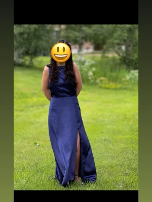 Marinblå långklänning med slits - Elegant marinblå långklänning i ett glansigt material med hög slits framtill och öppen rygg. Klänningen har en mjukt draperad rygg och är ärmlös, vilket ger en stilren och festlig look.