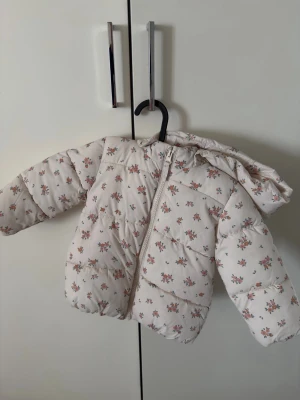 Blommig vit pufferjacka H&M baby - Supersöt vit pufferjacka från H&M med små rosa och gröna blommor. Jackan har huva, dragkedja framtill och mjukt fleecefoder på insidan. Passar barn 9-12 månader. Aldrig använd.