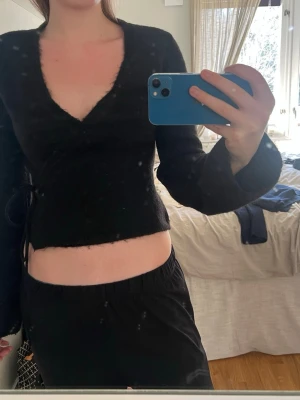 Svart omlottopp med v-ringning - Säljer en svart omlottopp med djup v-ringning och långa ärmar. Tröjan har en mjuk, lite luddig yta och knytdetalj i sidan. Passar perfekt till både jeans och kjol för en trendig look.