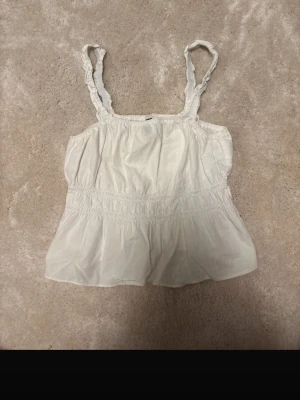 Vit linnetopp med rynkade axelband - Säljer en söt vit linnetopp med rynkade axelband och smockdetaljer runt midjan. Toppen är ärmlös och har en luftig passform som är perfekt för varma dagar. Materialet känns mjukt och lätt mot huden.