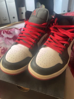 Air Jordan 1 Mid svart/vit/röd - Snygga Air Jordan 1 Mid sneakers i svart, vitt och rött med klassisk Nike Swoosh på sidan. Skorna har röda skosnören, perforerad tåbox och ikoniska Air Jordan-loggan på sidan. Ovandelen är i läder och sulan är vit med röd yttersula.