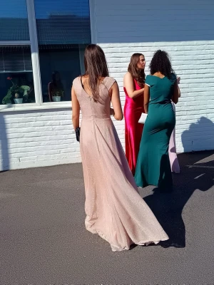 Lång glittrig champagneklänning - Elegant långklänning i champagnefärgad ton med glittrigt tyg och v-ringad rygg. Klänningen har breda axelband och ett svepande fall som ger en lyxig känsla. Perfekt för bal eller festliga tillfällen. Den är använd den en gång och har inga defekter förutom lite slitningar där nere. Har dessutom sytt vid bröstkorgen men det är ingenting man inte kan ändra på om man skulle vilja.  Originalpris 1300 SEK