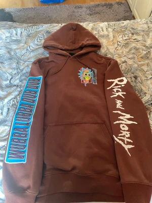 Rick and Morty brun hoodie - Säljer en brun hoodie med stor färgglad Rick and Morty-grafik på ryggen och mindre tryck på bröstet. Ena ärmen har texten 'WUBBA LUBBA DUB DUB' i blått och den andra 'Rick and Morty' i vitt. Klassisk känguruficka och huva. Perfekt för dig som älskar serien!