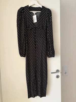 Svart prickig långklänning  - Svart långklänning från H&M med vita prickar, v-ringad hals och långa puffärmar. Klänningen har knappar framtill och är tillverkad i ett mjukt, lätt material med 55% viskos. Storlek XXS med prislappen kvar. 