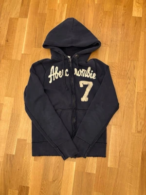 Marinblå hoodie Abercrombie & Fitch - Snygg marinblå hoodie från Abercrombie & Fitch med dragkedja, huva och stora broderade bokstäver samt siffran 7 på bröstet. Mjuk insida och kängurufickor framtill. Perfekt för en avslappnad och trendig stil.