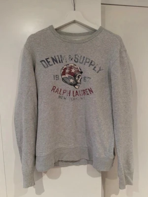 Grå sweatshirt från Ralph Lauren - Grå sweatshirt från Ralph Lauren Denim & Supply med tryck av en dödskalle i hjälm och texten '1967 Ralph Lauren New York'. Rund halsringning och ribbade muddar vid ärmar och nederkant. Hör gärna av dig vid frågor!