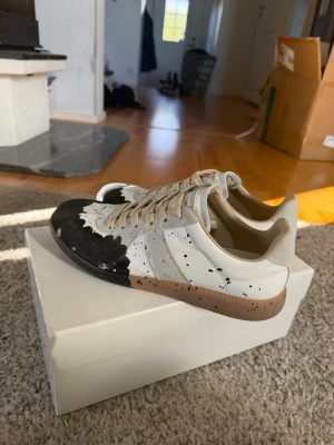 Maison Margiela skor  - Maison Margiela sneakers i vitt och beige läder och mocka med svart färgdetalj framtill och färgstänk över hela skon. Klassisk låg modell med beige snörning och gummisula. Unik design som sticker ut och passar perfekt till streetwear.