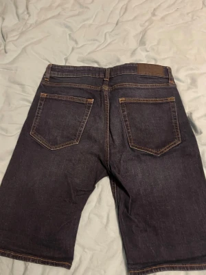 Mörkblå jeansshorts från Dressmann - Snygga mörkblå jeansshorts från Dressmann med klassisk femficksdesign och kontrastsömmar. Shortsen har bälteshällor och knappgylf. Perfekta för en avslappnad stil under varmare dagar.