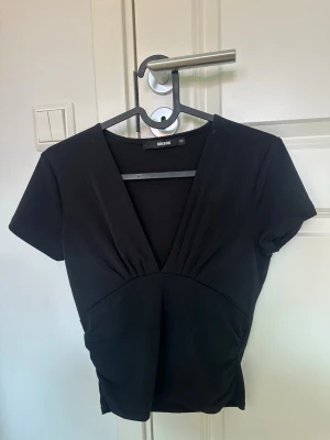 Svart v-ringad topp från BikBok - Svart topp från BikBok i storlek XS med djup v-ringning och korta ärmar. Snygg passform som framhäver midjan och stilren design.  