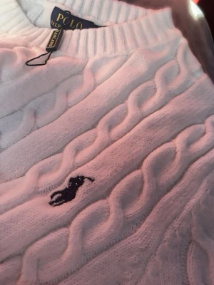  Polo Ralph Lauren - Vit kabelstickad tröja från Polo Ralph Lauren med klassisk rund halsringning och broderad logotyp i mörkblått på bröstet. Tröjan är gjord i mjukt stickat material och har ribbade muddar vid hals och ärmslut. Perfekt för en clean och stilren look.