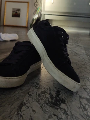 Mörkblå sneakers från Les Deux - Snygga mörkblå sneakers från Les Deux med ovandel i mocka och vita sulor. Skorna har mörkblå skosnören och diskreta detaljer i rött och vitt på hälen. Perfekta för en stilren och avslappnad look.