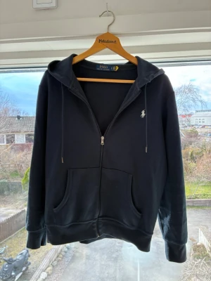 Svart zip hoodie från Polo Ralph Lauren - Säljer en svart hoodie med dragkedja från Polo Ralph Lauren. Klassisk design med huva, fickor framtill och den ikoniska vita logon broderad på bröstet. Mjuk bomullsblandning och ribbade muddar vid ärmar och nederkant. Perfekt för en avslappnad stil.