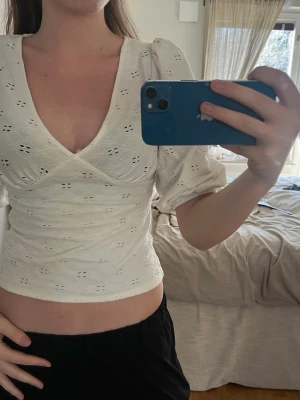 Vit omlott blus med puffärm - Säljer en vit blus med omlott-design och v-ringning. Blusen har puffiga korta ärmar och är tillverkad i bomull med broderade hålmönster. Den är croppad och har en feminin, figurnära passform som framhäver midjan.