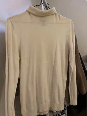 Beige polotröja i stickat material - Klassisk beige stickad polotröja med lång ärm och ribbad krage. Tröjan har en enkel och stilren design som passar till det mesta. Mjuk och skön kvalitet, perfekt för lager på lager under kyliga dagar.