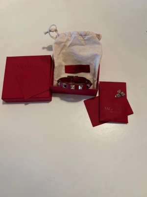 Rött Valentino armband - Säljer detta feta valentino armbandet då det inte kommer till så mycke användning längre alla tillbehör är med som två extra nitar osv. Lite slitage vid ändan men syns inte när man har det på