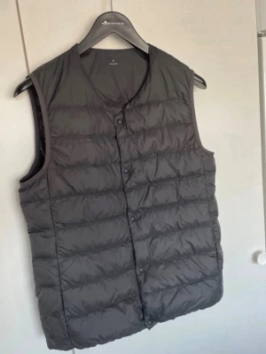 Svart dunväst från Muji - Stilren svart dunväst från Muji med knäppning framtill och klassisk quiltad design. Västen har en enkel, rak passform och är tillverkad i lätt och smidigt material med dunfyllning. Perfekt att slänga över en hoodie eller tröja för extra värme.