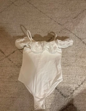 Vit off-shoulder body med volang - Säljer en vit body med smala axelband och off-shoulder volangdetaljer. Bodyn har tryckknappar i grenen och är tillverkad i ett mjukt, stretchigt tyg. Perfekt för dig som gillar en romantisk och trendig stil.