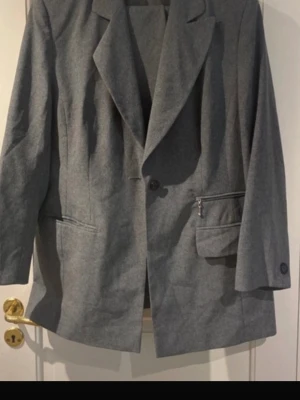 Kostym Grå oversized kavaj med fickor och byxor  - Stilren grå kavaj i oversized modell med klassisk krage och två framfickor, varav en med dragkedja. Kavajen har enkelknäppning och knappar vid ärmsluten. Perfekt för en trendig och avslappnad look.