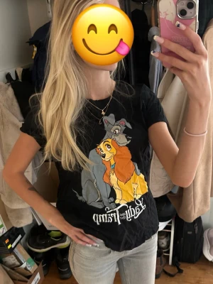 Svart Disney Lady & Lufsen T-shirt - Säljer en svart T-shirt från FB Sister x Disney med tryck av Lady & Lufsen framtill. T-shirten har korta ärmar, rund hals och är i mjuk bomull. Perfekt för dig som älskar Disney och vill ha en chill och snygg look.