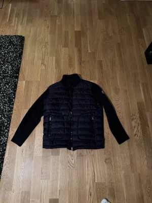 Mörkblå Moncler cardiganjacka - Snygg mörkblå cardiganjacka från Moncler med quiltad front och stickade ärmar. Jackan har två dragkedjeförsedda fickor och Moncler-logga på ärmen. Perfekt för lager-på-lager och har en hög krage för extra stil.