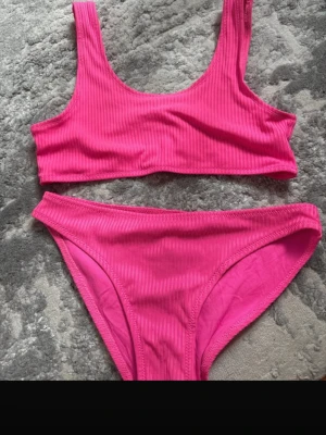 Rosa ribbad bikini - Säljer en snygg rosa bikini med ribbad struktur. Bikinin har en sportig topp med breda axelband och matchande trosor med klassisk passform. Perfekt för stranden eller poolen i sommar!