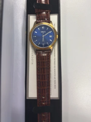 Sirius klocka med blå urtavla - Stilren klocka från Sirius med blå urtavla, guldfärgat boett och klassiskt brunt armband i skinn med krokodilpräglat mönster. Urtavlan har romerska siffror och datumvisning. Perfekt accessoar för dig som gillar tidlös design.