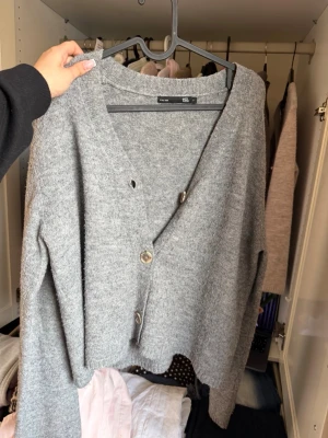 Grå kofta från 157 med knappar - Mysig grå kofta från 157 med v-ringning och tre knappar framtill. Koftan har en avslappnad passform och långa ärmar, perfekt att slänga över en t-shirt eller topp. Enkel och stilren design som passar till det mesta.