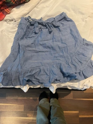 Blå volangkjol med knytband - Supersöt blå kortkjol med volangkant och knytband i midjan. Kjolen har en avslappnad passform och är gjord i ett lätt och luftigt bomullsliknande material, perfekt för varma dagar. Enkel att styla med både t-shirt och linne.