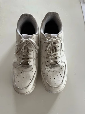 Vita Nike Air Force 1 sneakers - Klassiska vita Nike Air Force 1 sneakers i lågt utförande. Skorna har en stilren design med perforerad tå, vita skosnören och den ikoniska swoosh-loggan på sidan. Ovandelen är i läder och sulan är tjock och greppvänlig. Perfekta för en clean och tidlös look.