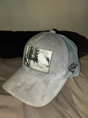 Grisch keps Ca'put headwear - Säljer en grå keps från Ca'put headwear med mesh på sidorna och ett svartvitt stadsmotiv broderat framtill. Kepsen har böjd skärm och justerbar baksida. Perfekt för dig som gillar streetwear och unika detaljer.