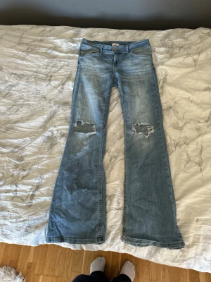 Ljusblå bootcut jeans med slitningar - Säljer ett par ljusblå bootcut jeans med slitna hål på båda knäna och lätt tvättad look. Jeansen har klassisk femficksmodell och vida ben. Perfekta för dig som gillar en avslappnad och trendig stil.