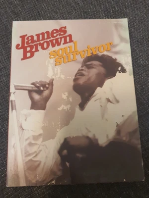 James Brown Soul Survivor dvd - En dvd med titeln 'James Brown Soul Survivor' som handlar om James Browns liv och musikaliska resa. Omslaget har ett foto av James Brown vid en mikrofon och text i rött och gult. Dvdn är tryckt på papper och har ett färgstarkt omslag.