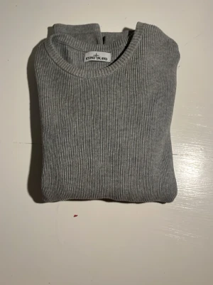 Grå ribbstickad tröja Stone Island - Snygg grå ribbstickad tröja från Stone Island med rund halsringning och lång ärm. Tröjan har en diskret logga på ärmen och är tillverkad i ett mjukt, stretchigt material som sitter skönt på kroppen. Perfekt för dig som gillar stilrena och klassiska plagg.