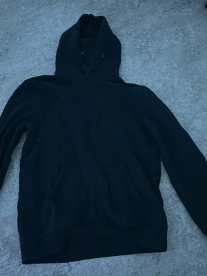 Svart basic hoodie med huva - En enkel svart hoodie med klassisk huva och känguruficka framtill. Hoodien har långa ärmar och ribbade muddar vid ärmslut och nederkant. Perfekt för en avslappnad och stilren look.