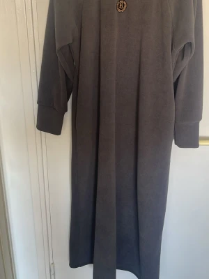 Mörkgrå morgonrock med emblem - Mysig och lång morgonrock i mörkgrå färg med ett litet emblem på bröstet. Morgonrocken har långa ärmar och ett klassiskt bälte i midjan. Perfekt för att chilla hemma och hålla sig varm.