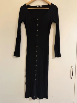 Svart ribbad långklänning med knappar - Svart långklänning med ribbad struktur och knappar längs hela framsidan. Klänningen har lång ärm och djup rundringning. Perfekt för dig som gillar en stilren och enkel look.